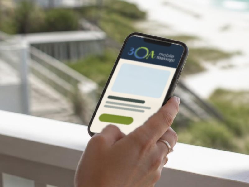 A hand holding a phone showing the 30A Mobile Massage site on a beach-rental balcony